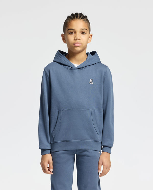 A child with braided hair is wearing the KIDS LEVY ESSENTIAL FLEECE HOODIE by Psycho Bunny and matching fleece sweatpants, standing against a plain white background.