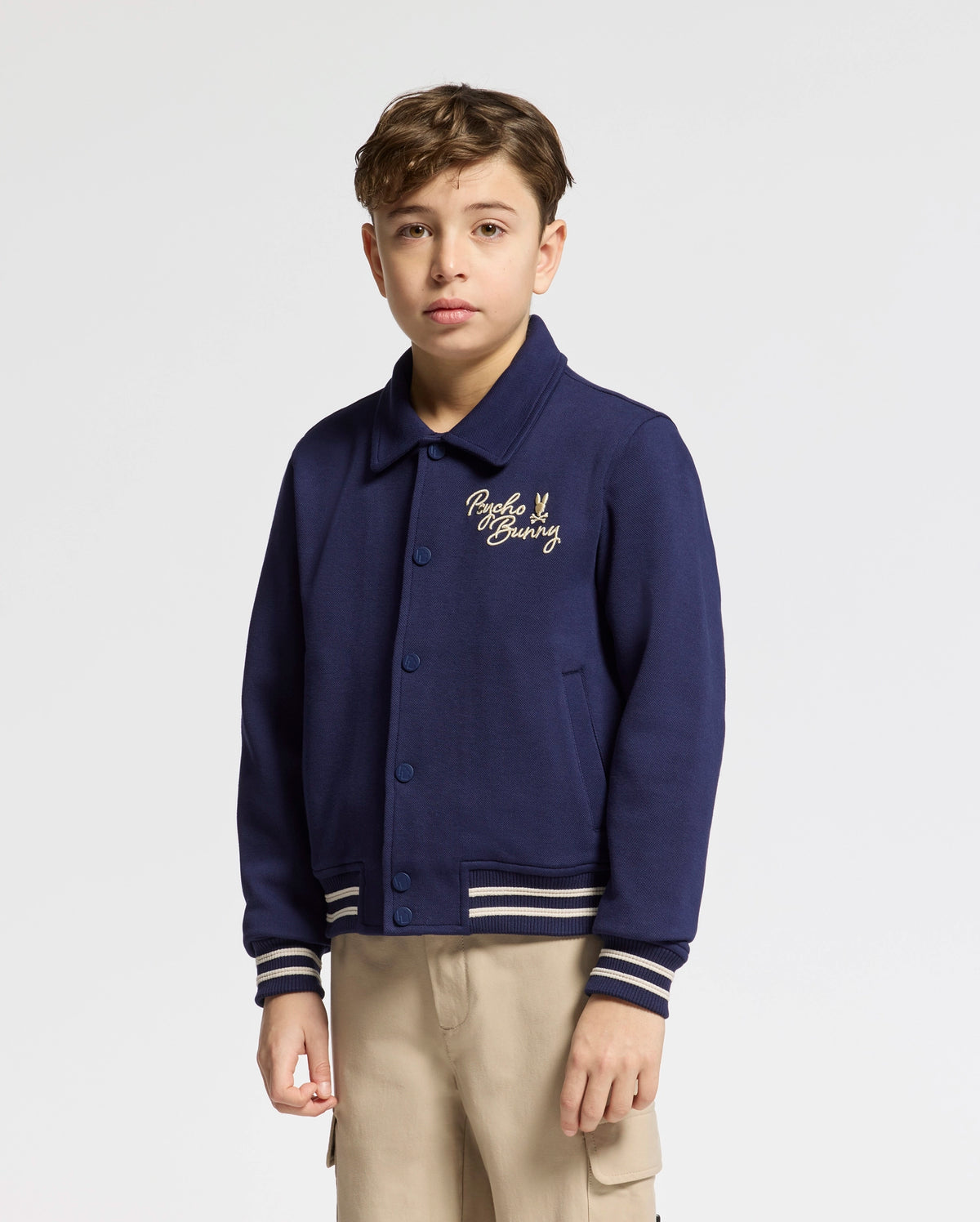 A young boy in a Psycho Bunny KIDS DESERT COACH BOMBER JACKET - B0J428F200, Evening Blue with chest embroidery, stands against a white backdrop. The Double Knit Jacquard jacket features a collar and striped cuffs, perfectly complementing his light-colored pants.
