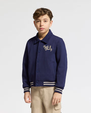 A young boy in a Psycho Bunny KIDS DESERT COACH BOMBER JACKET - B0J428F200, Evening Blue with chest embroidery, stands against a white backdrop. The Double Knit Jacquard jacket features a collar and striped cuffs, perfectly complementing his light-colored pants.