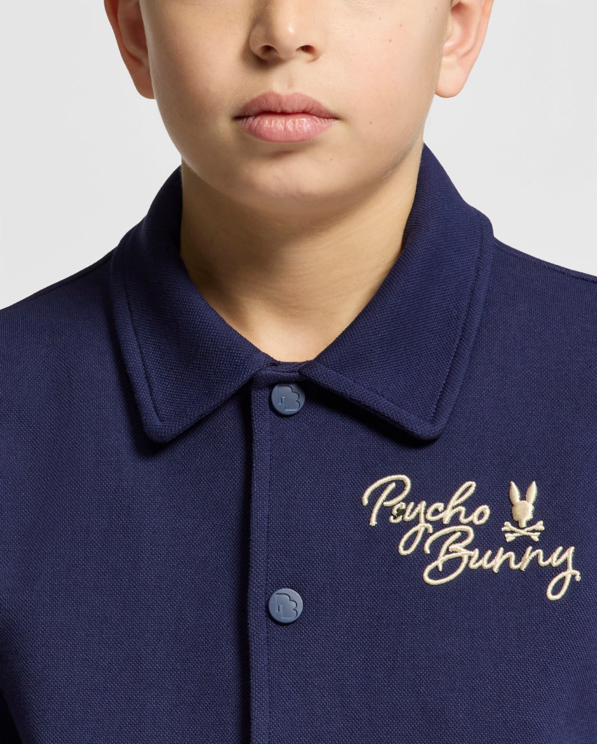 Wearing an Evening Blue collared shirt featuring "Psycho Bunny" embroidery and a small beige bunny logo on the chest, reminiscent of the elegance found in a Double Knit Jacquard design, stands out against a plain light gray backdrop.