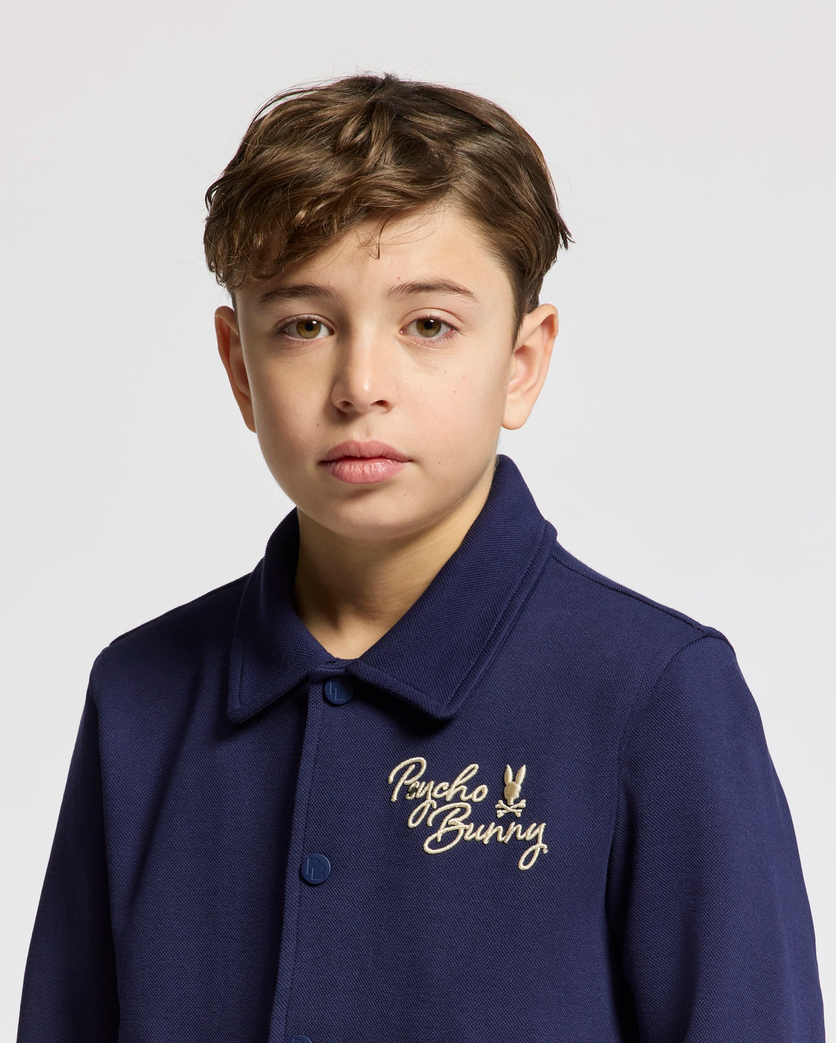 A young person with short brown hair wears the KIDS DESERT COACH BOMBER JACKET - B0J428F200 by Psycho Bunny, featuring elegant embroidery on the chest. The Evening Blue hue of the jacket pops against a plain white background for a stylish statement.