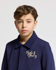 A young person with short brown hair wears the KIDS DESERT COACH BOMBER JACKET - B0J428F200 by Psycho Bunny, featuring elegant embroidery on the chest. The Evening Blue hue of the jacket pops against a plain white background for a stylish statement.