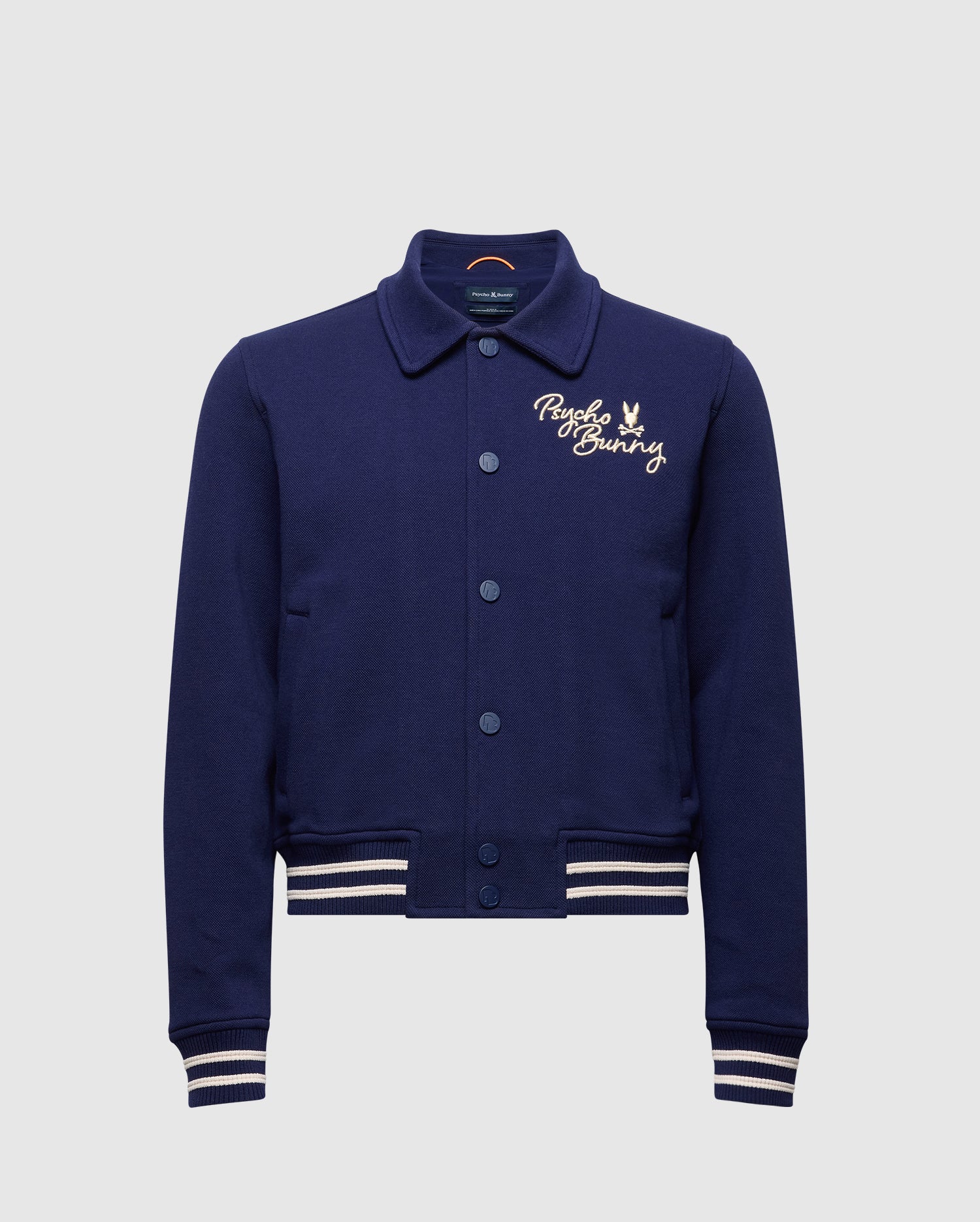 The KIDS DESERT COACH BOMBER JACKET - B0J428F200 by Psycho Bunny is an evening blue varsity-style jacket with ribbed cuffs and hem featuring white stripes, a button-up front showcasing "Psycho Bunny" embroidered in light beige on the left chest, and a small bunny logo.
