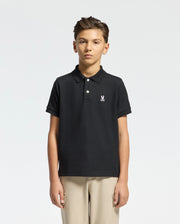 A young boy wears a Psycho Bunny Kids Classic Pique Polo Shirt (B0K001CRPC) featuring a white embroidered logo on the left chest, paired with beige trousers. Perfect for back-to-school outfits, he stands confidently against a plain white background.