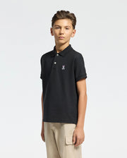 A young boy stands against a plain background, wearing a Psycho Bunny KIDS CLASSIC PIQUE POLO SHIRT and khaki cargo pants—ideal for back to school. He has short hair and a neutral expression.