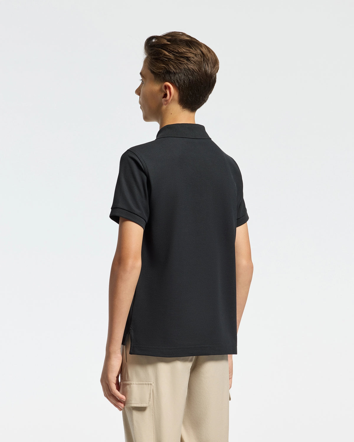 A person with short brown hair is viewed from the back in a Psycho Bunny KIDS CLASSIC PIQUE POLO SHIRT - B0K001CRPC, paired with beige pants against a simple white backdrop, highlighting their timeless look.
