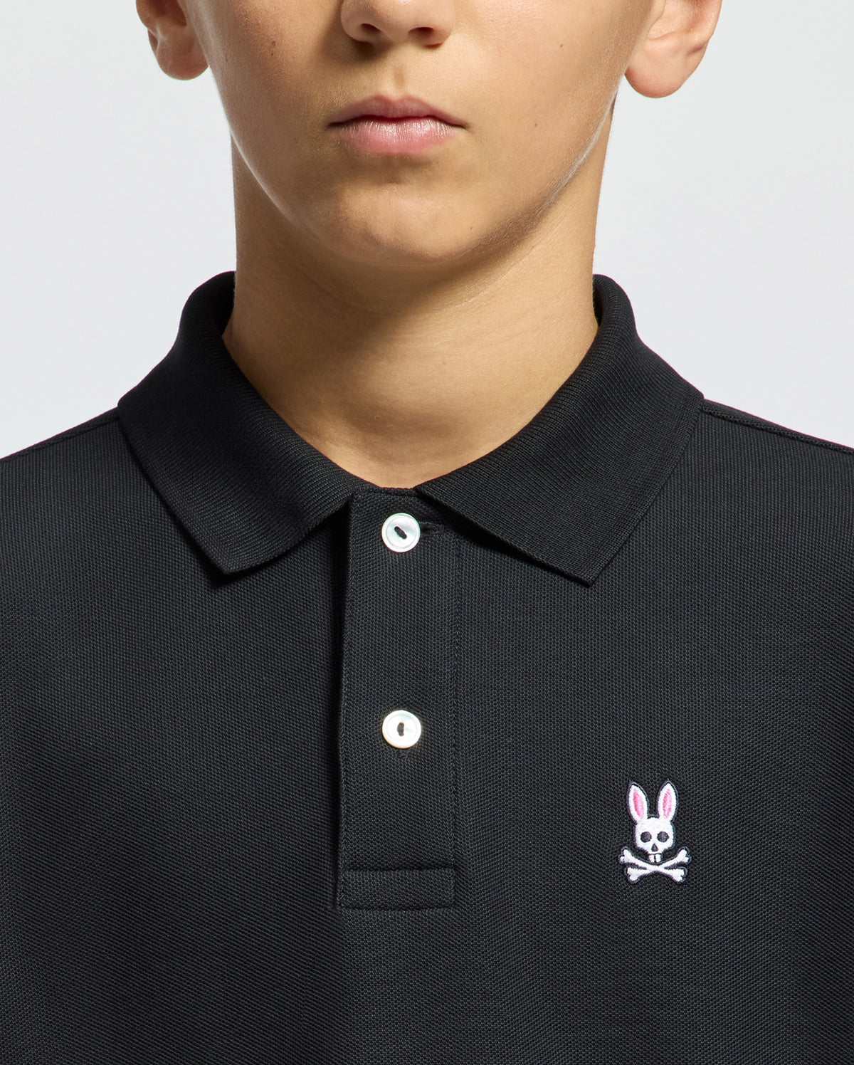 A person wears a Psycho Bunny Kids Classic Pique Polo Shirt (B0K001CRPC) in black, featuring a collar and buttons. It displays a small embroidered logo of a pink-eared bunny with skull and crossbones on the chest, ideal for back-to-school outfits, against a plain light gray background.