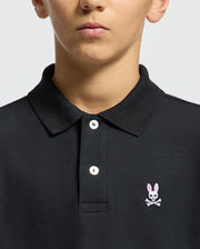A person wears a Psycho Bunny Kids Classic Pique Polo Shirt (B0K001CRPC) in black, featuring a collar and buttons. It displays a small embroidered logo of a pink-eared bunny with skull and crossbones on the chest, ideal for back-to-school outfits, against a plain light gray background.