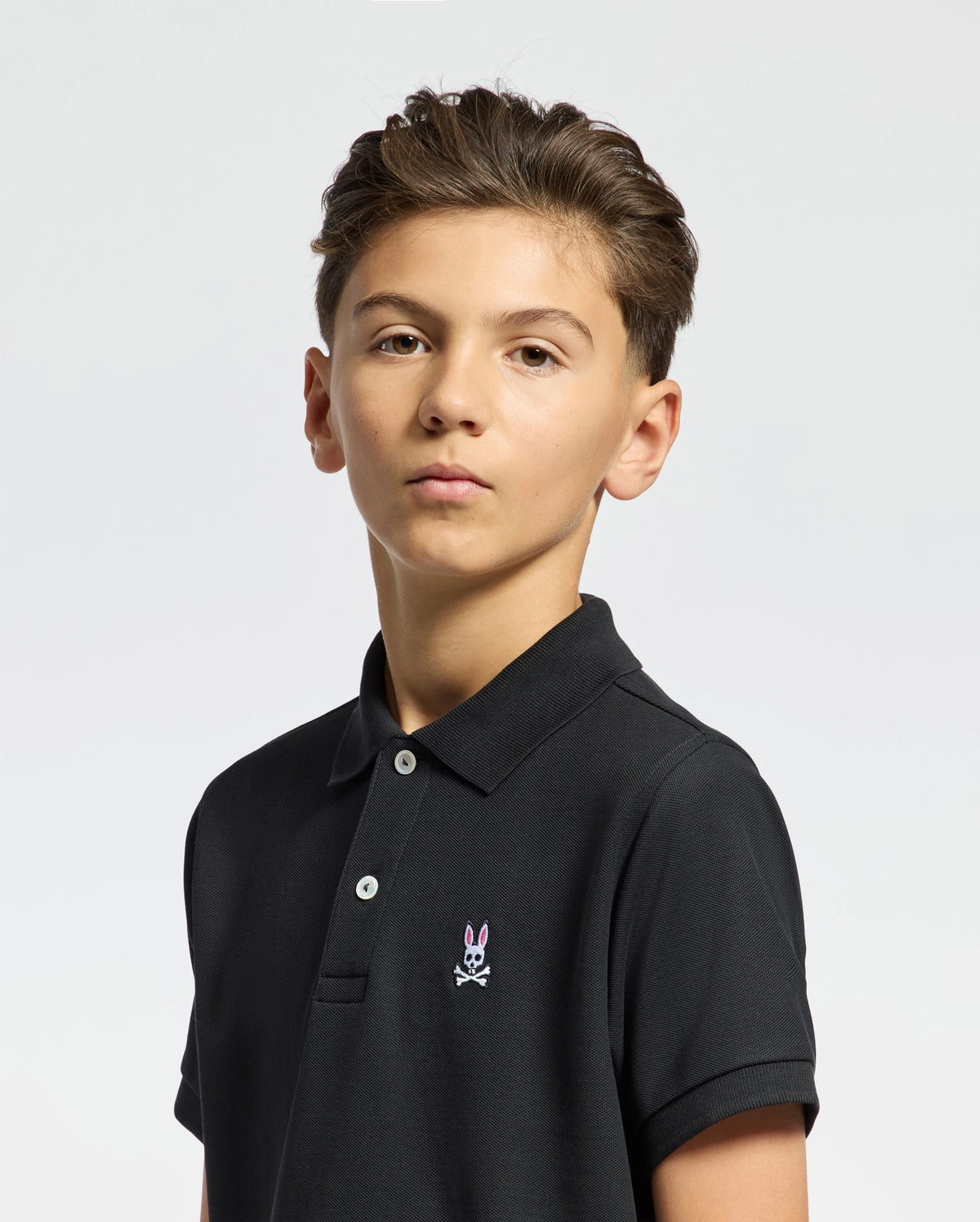 A young person with short brown hair models Psycho Bunny's KIDS CLASSIC PIQUE POLO SHIRT - B0K001CRPC. The classic black polo, featuring an embroidered logo on the chest, is ideal for back-to-school outfits. The simple yet stylish look stands out against a plain, light-colored background.