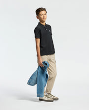 Against a plain background, a person wears the Psycho Bunny KIDS CLASSIC PIQUE POLO SHIRT in black, beige pants, and sneakers. They hold a denim jacket by their side, creating an effortlessly stylish back-to-school look.