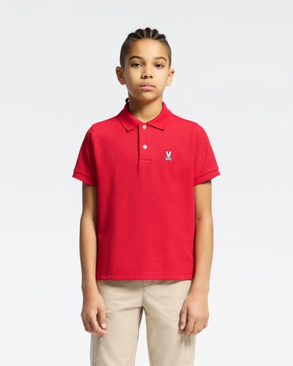 A young person with braided hair wears a red Psycho Bunny KIDS CLASSIC PIQUE POLO SHIRT (B0K001CRPC) featuring a small rabbit emblem on the chest, paired with beige pants, set against a white background—ideal for back-to-school style.