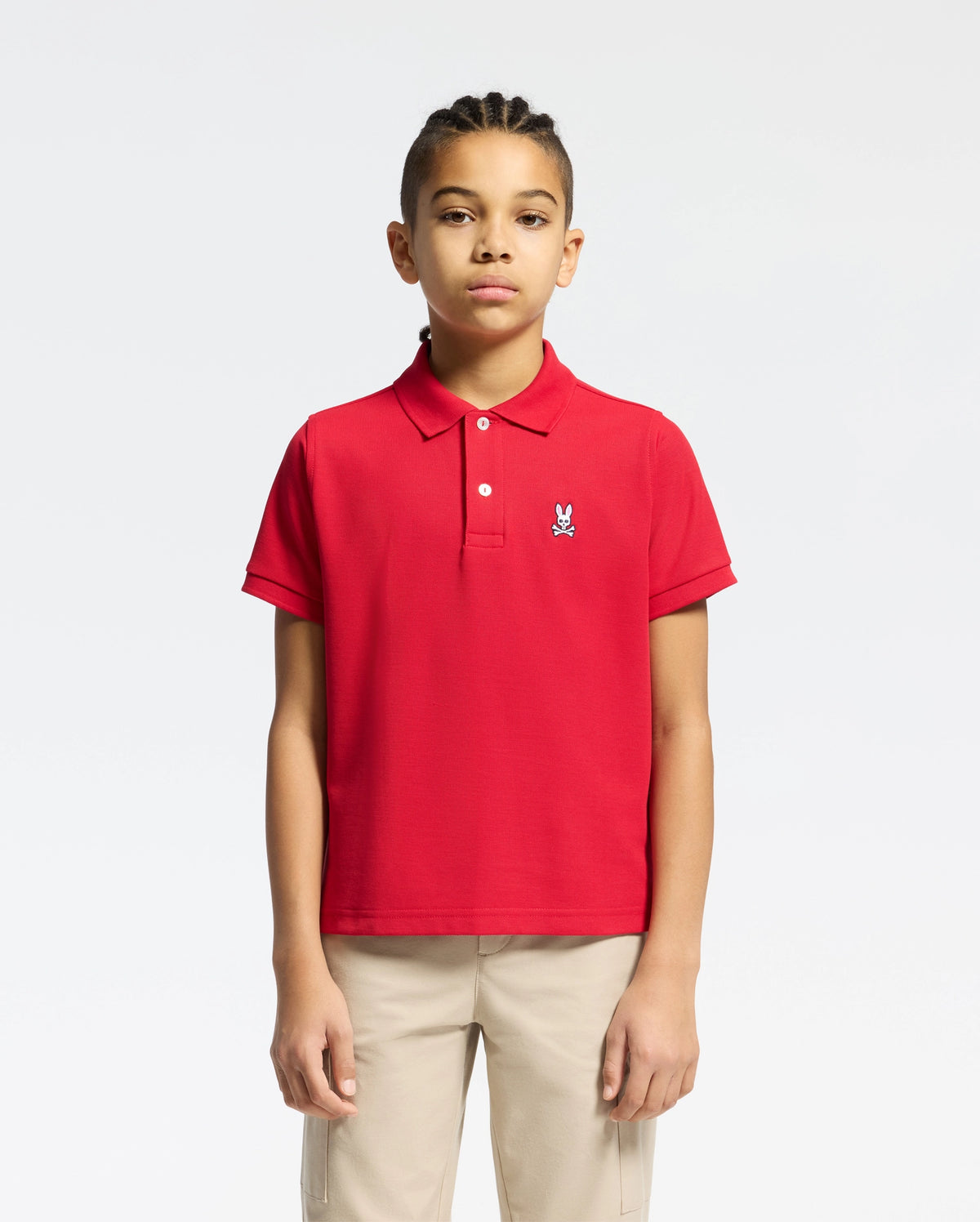 A young person with braided hair wears a red Psycho Bunny KIDS CLASSIC PIQUE POLO SHIRT (B0K001CRPC) featuring a small rabbit emblem on the chest, paired with beige pants, set against a white background—ideal for back-to-school style.