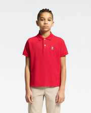 A young person with braided hair wears a red Psycho Bunny KIDS CLASSIC PIQUE POLO SHIRT (B0K001CRPC) featuring a small rabbit emblem on the chest, paired with beige pants, set against a white background—ideal for back-to-school style.