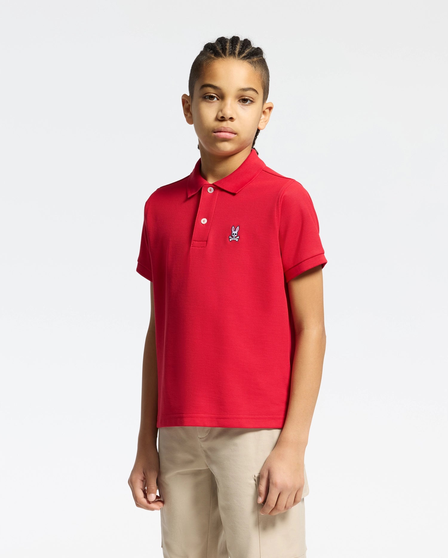 A young person with braided hair is wearing a Psycho Bunny KIDS CLASSIC PIQUE POLO SHIRT in bold red and beige pants, ideal for back-to-school outfits. The shirt features a small logo on the left against a plain white background.