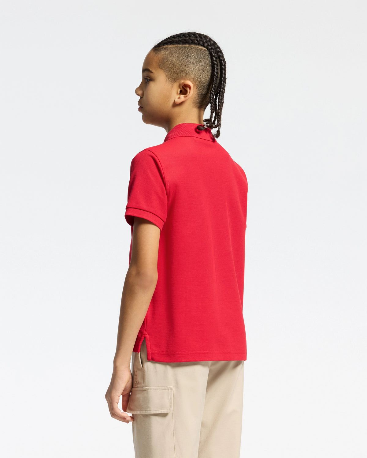 With braided hair, a person stands sideways in a red Psycho Bunny kids' classic pique polo shirt (B0K001CRPC) and beige pants with large pockets. The plain white background enhances this perfect back-to-school outfit.
