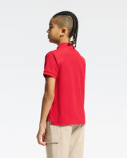 With braided hair, a person stands sideways in a red Psycho Bunny kids' classic pique polo shirt (B0K001CRPC) and beige pants with large pockets. The plain white background enhances this perfect back-to-school outfit.