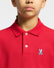The Psycho Bunny KIDS CLASSIC PIQUE POLO SHIRT (B0K001CRPC) features a red color, embroidered rabbit and crossed bones logo on the chest, and a neatly buttoned collar. It's ideal for creating a sharp back-to-school look against a plain white background.