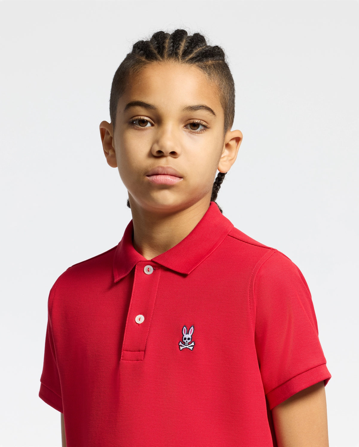 A child with braided hair wears a Psycho Bunny KIDS CLASSIC PIQUE POLO SHIRT in classic red, featuring a rabbit emblem. This outfit is perfect for back-to-school, set against a solid white backdrop.