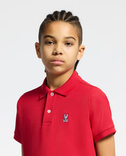 A child with braided hair wears a Psycho Bunny KIDS CLASSIC PIQUE POLO SHIRT in classic red, featuring a rabbit emblem. This outfit is perfect for back-to-school, set against a solid white backdrop.