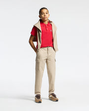 A young person, confidently posing with hands in pockets, sports a Psycho Bunny KIDS CLASSIC PIQUE POLO SHIRT in red, beige cargo pants, a beige hoodie draped over their shoulders, braided hair, and brown sneakers. The perfect back-to-school look against a plain white background.