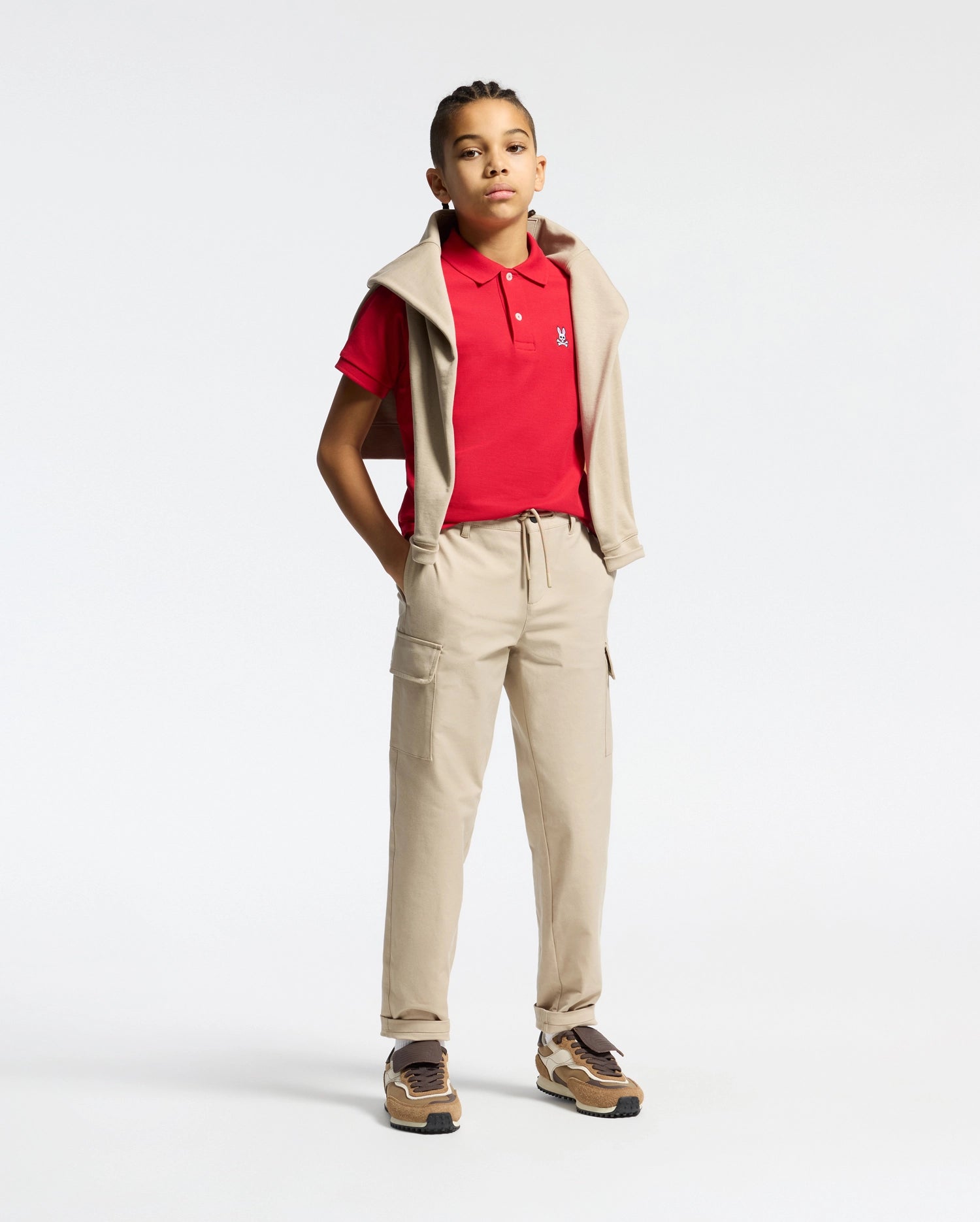 A young person, confidently posing with hands in pockets, sports a Psycho Bunny KIDS CLASSIC PIQUE POLO SHIRT in red, beige cargo pants, a beige hoodie draped over their shoulders, braided hair, and brown sneakers. The perfect back-to-school look against a plain white background.