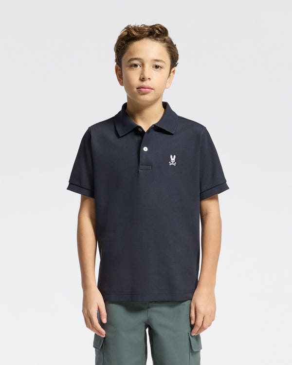 A young individual wearing the Psycho Bunny Kids Classic Pique Polo Shirt (B0K001CRPC), displaying a small chest emblem, stands against a white backdrop. With short, curly hair and a confident look, they capture the stylish essence of back-to-school fashion.