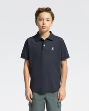 A young individual wearing the Psycho Bunny Kids Classic Pique Polo Shirt (B0K001CRPC), displaying a small chest emblem, stands against a white backdrop. With short, curly hair and a confident look, they capture the stylish essence of back-to-school fashion.