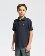 A brown-haired boy models the Psycho Bunny KIDS CLASSIC PIQUE POLO SHIRT - B0K001CRPC in navy, highlighted by a small white logo on the left chest. His green cargo pants complete this standout back-to-school look against a plain white backdrop.