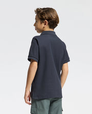 A young boy with short brown hair, wearing a Psycho Bunny Kids Classic Pique Polo Shirt and green cargo shorts, stands facing away against a light gray background. His classic back-to-school look exudes both style and comfort.