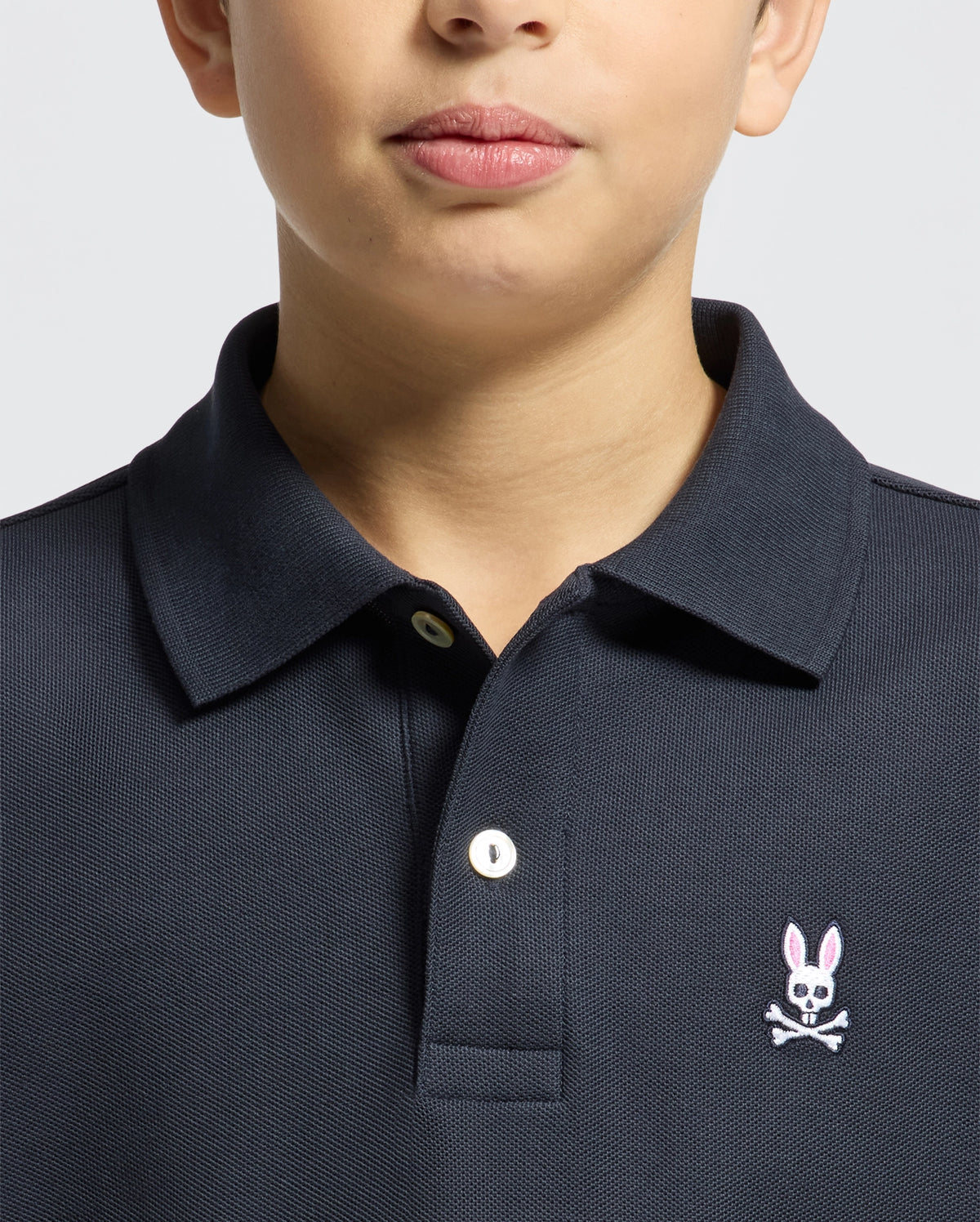 A child models the Psycho Bunny Kids Classic Pique Polo Shirt (B0K001CRPC) with a pink rabbit and crossbones chest logo against a plain gray background. Only the lower face is visible, making this chic polo perfect for back-to-school outfits.
