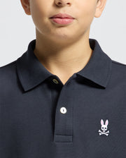 A child models the Psycho Bunny Kids Classic Pique Polo Shirt (B0K001CRPC) with a pink rabbit and crossbones chest logo against a plain gray background. Only the lower face is visible, making this chic polo perfect for back-to-school outfits.
