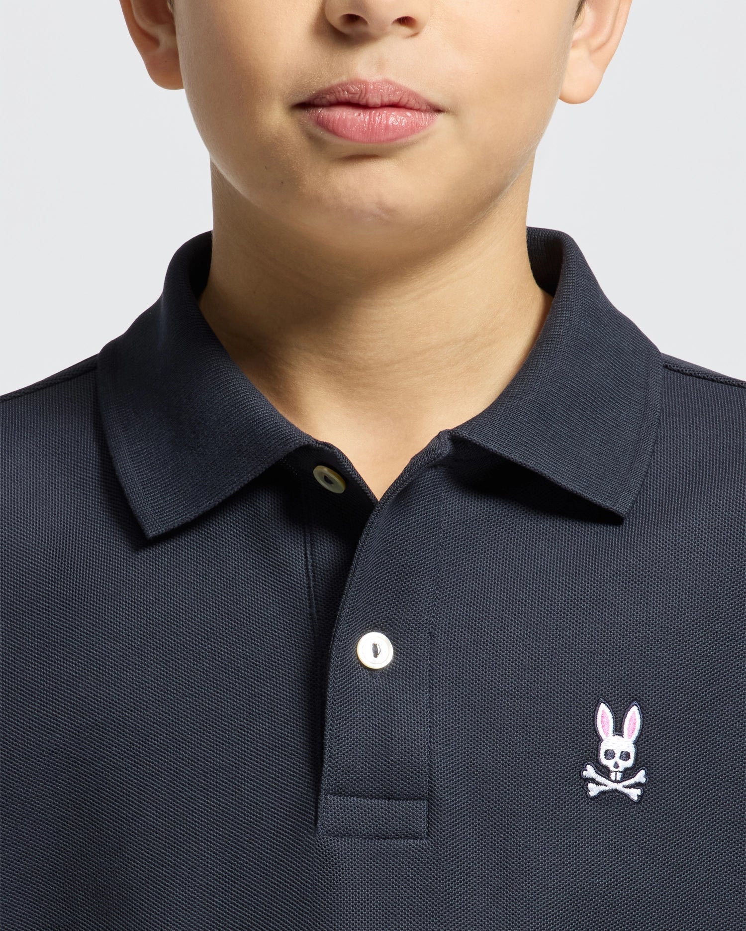 A child models the Psycho Bunny Kids Classic Pique Polo Shirt (B0K001CRPC) with a pink rabbit and crossbones chest logo against a plain gray background. Only the lower face is visible, making this chic polo perfect for back-to-school outfits.