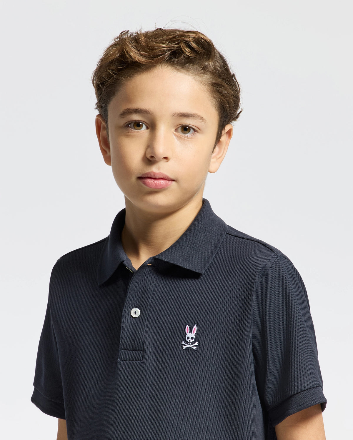 A young boy with short, light brown hair wears the KIDS CLASSIC PIQUE POLO SHIRT - B0K001CRPC by Psycho Bunny. This classic navy polo, featuring a small bunny logo, is an ideal back-to-school outfit. He stands with a neutral expression against a plain white background.