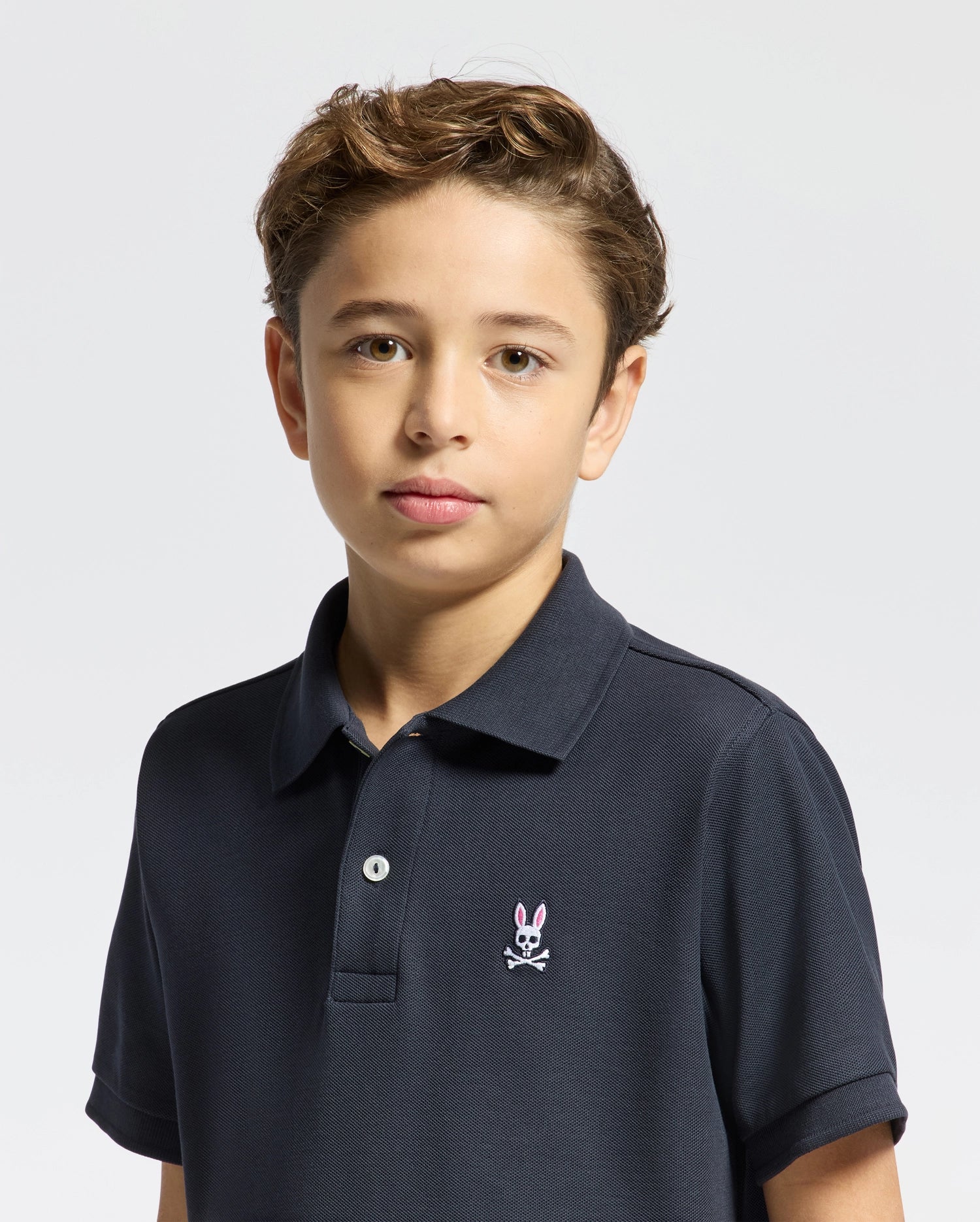 A young boy with short, light brown hair wears the KIDS CLASSIC PIQUE POLO SHIRT - B0K001CRPC by Psycho Bunny. This classic navy polo, featuring a small bunny logo, is an ideal back-to-school outfit. He stands with a neutral expression against a plain white background.