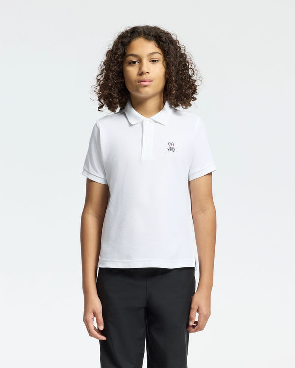 With curly hair, a person models the Psycho Bunny Kids Classic Pique Polo Shirt (white) against a plain white background, paired with black pants. Ideal for back-to-school, it features a subtle left-side graphic.