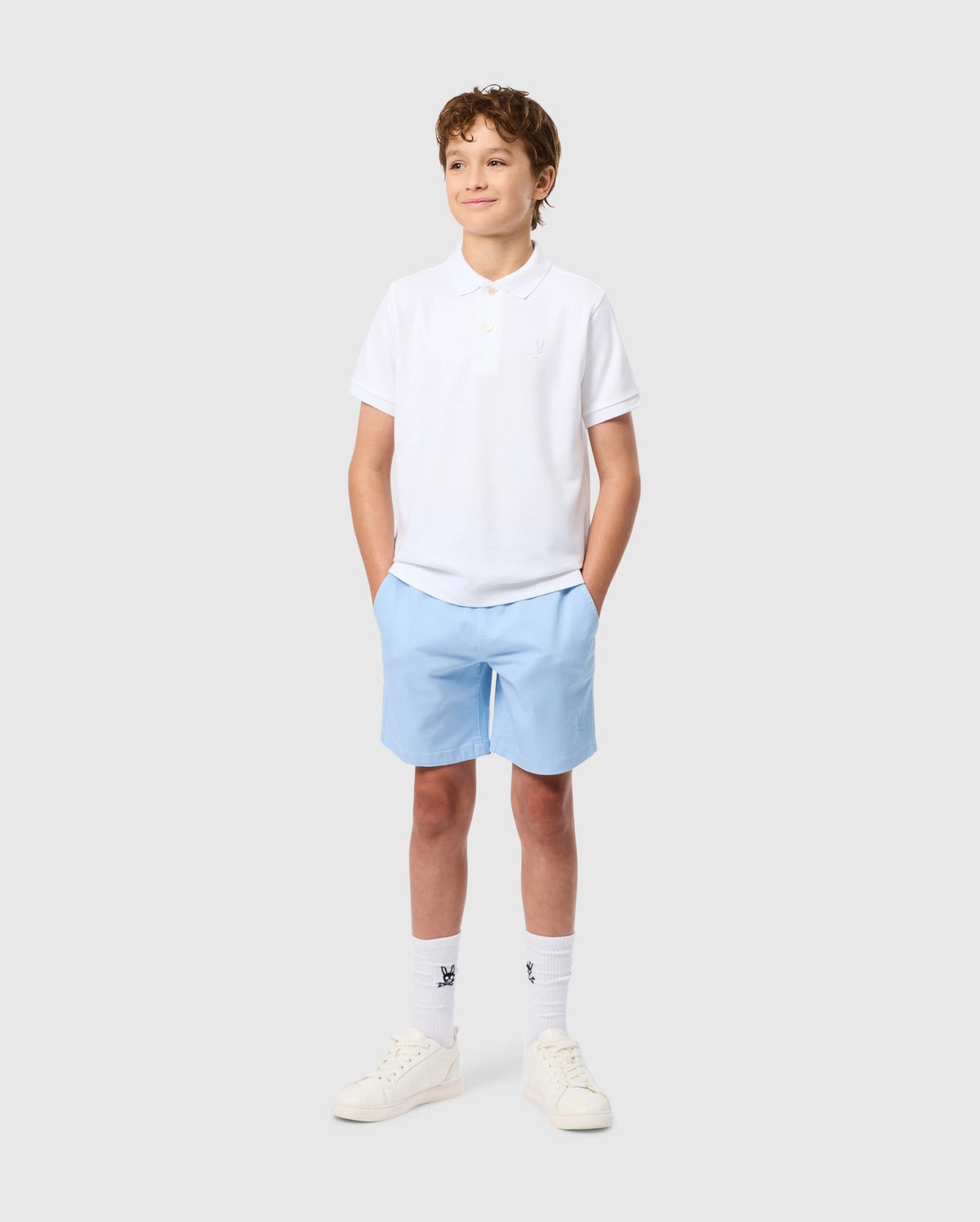 A young boy smiles with hands in pockets, wearing the Psycho Bunny KIDS CLASSIC PIQUE POLO SHIRT made from soft Pima cotton. His outfit features light blue shorts, white socks, and sneakers. An embroidered Bunny adorns his shirt on a plain gray background.