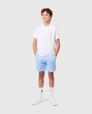 A young boy smiles with hands in pockets, wearing the Psycho Bunny KIDS CLASSIC PIQUE POLO SHIRT made from soft Pima cotton. His outfit features light blue shorts, white socks, and sneakers. An embroidered Bunny adorns his shirt on a plain gray background.