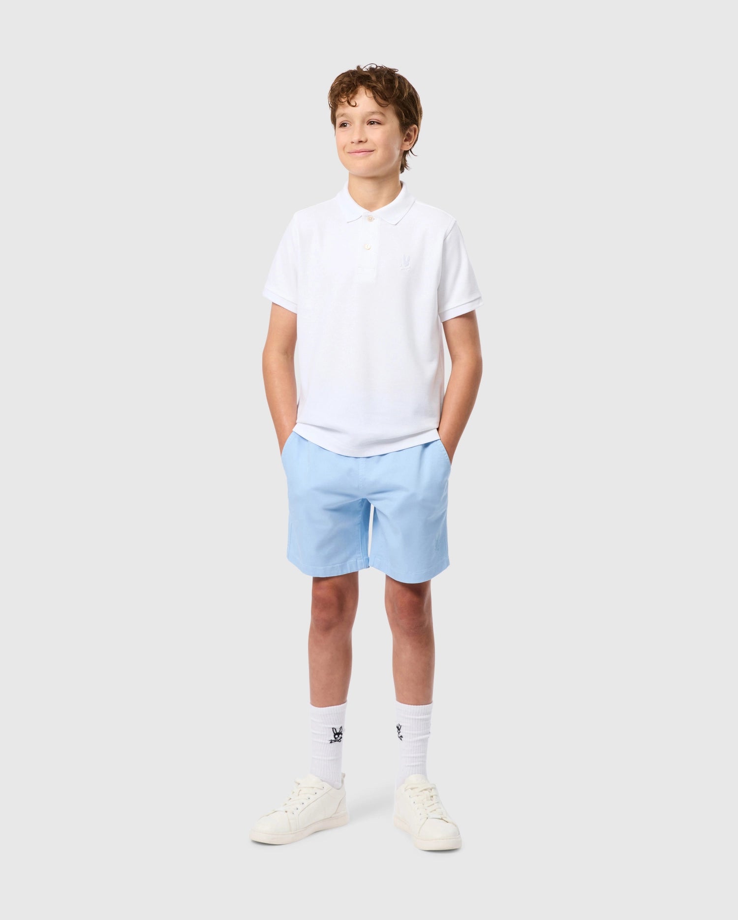 A young boy smiles with hands in pockets, wearing the Psycho Bunny KIDS CLASSIC PIQUE POLO SHIRT made from soft Pima cotton. His outfit features light blue shorts, white socks, and sneakers. An embroidered Bunny adorns his shirt on a plain gray background.
