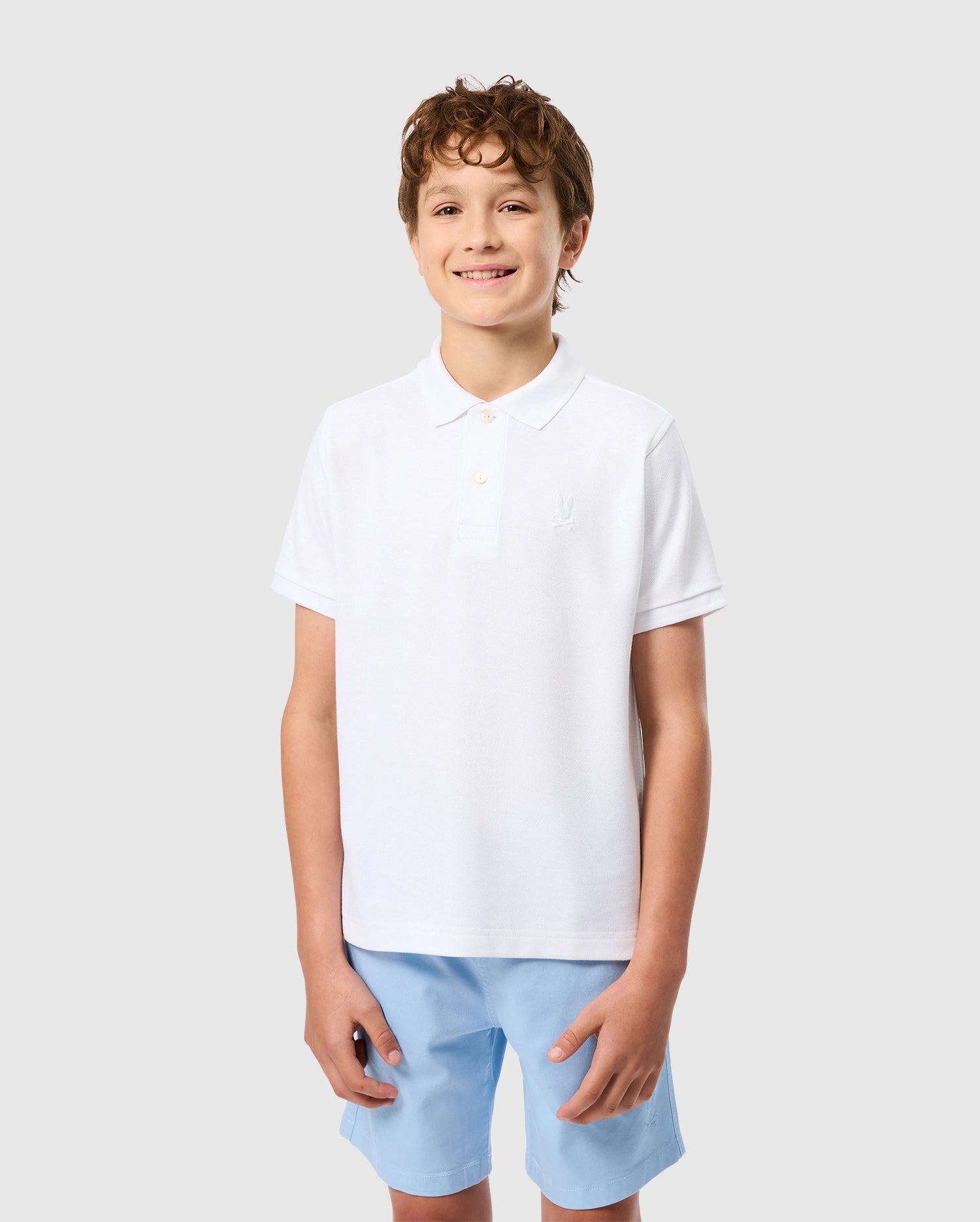 A smiling boy with curly hair wears Psycho Bunny's Kids Classic Pique Polo Shirt, crafted from soft Pima cotton, paired with light blue shorts. He stands against a plain light gray background, exuding charm and comfort.