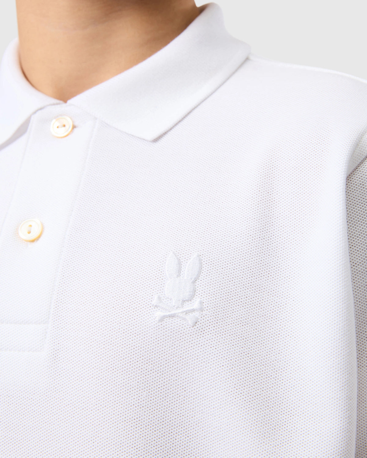 A person is wearing the Psycho Bunny KIDS CLASSIC PIQUE POLO SHIRT - B0K021AR00, made from luxurious Pima cotton. It features an embroidered Bunny and crossbones logo on the chest, textured fabric, a collar, and two visible buttons against a light gray background.