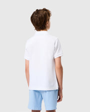 A brown-haired boy faces away from the camera wearing a Psycho Bunny KIDS CLASSIC PIQUE POLO SHIRT - B0K021AR00, stylishly paired with light blue shorts against a simple light gray backdrop.
