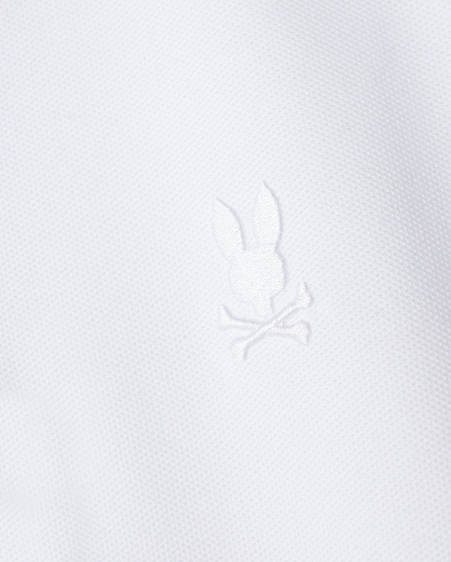 The Psycho Bunny Kids Classic Pique Polo Shirt (B0K021AR00) features a white fabric with a central embroidered Bunny motif above crossed bones, crafted in textured Pima cotton for a minimalist yet elegant design reminiscent of classic polo styles.