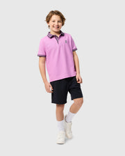 A smiling child stands against a plain background, wearing a light purple KIDS SOUTHPORT PIQUE POLO SHIRT - B0K263B200 by Psycho Bunny with a small embroidered logo, dark shorts, white socks, and white sneakers. The child has short, wavy hair and adopts a relaxed and friendly pose.