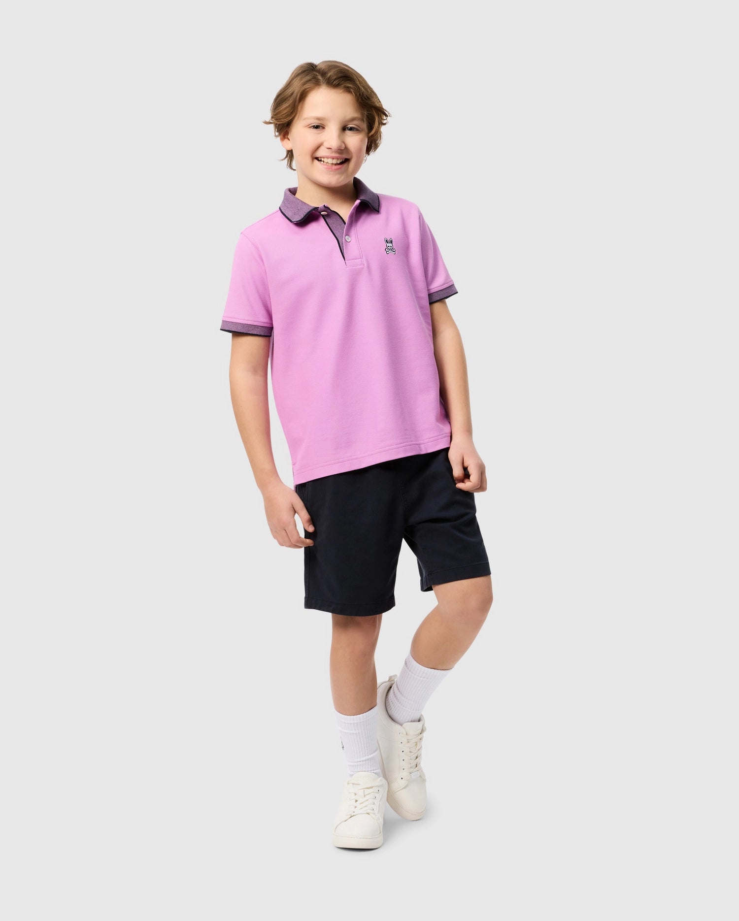 A smiling child stands against a plain background, wearing a light purple KIDS SOUTHPORT PIQUE POLO SHIRT - B0K263B200 by Psycho Bunny with a small embroidered logo, dark shorts, white socks, and white sneakers. The child has short, wavy hair and adopts a relaxed and friendly pose.