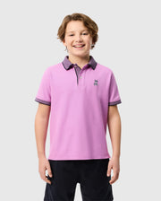 A smiling child with short brown hair is wearing a pink Psycho Bunny KIDS SOUTHPORT PIQUE POLO SHIRT - B0K263B200, featuring dark-colored collar and sleeve trim, a small embroidered logo on the chest, and crafted from soft Pima cotton. The child is standing against a plain light gray background.