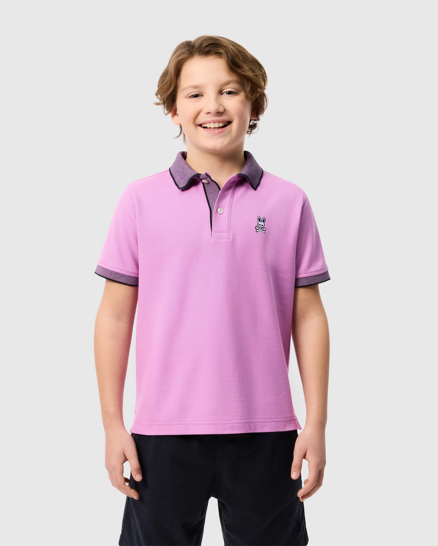A smiling child with short brown hair is wearing a pink Psycho Bunny KIDS SOUTHPORT PIQUE POLO SHIRT - B0K263B200, featuring dark-colored collar and sleeve trim, a small embroidered logo on the chest, and crafted from soft Pima cotton. The child is standing against a plain light gray background.