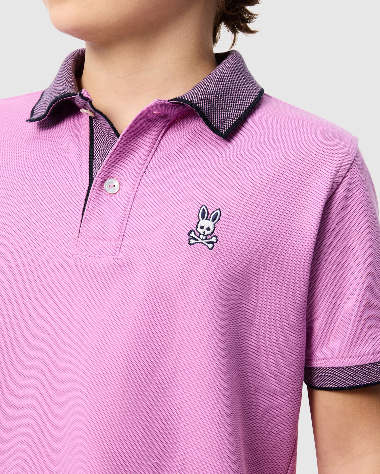 A person wearing a Psycho Bunny KIDS SOUTHPORT PIQUE POLO SHIRT - B0K263B200 in light purple with a black and white collar. The shirt, made from luxurious Pima cotton, features a small embroidered logo of a bunny above crossed bones on the left chest. The background is plain and light-colored.