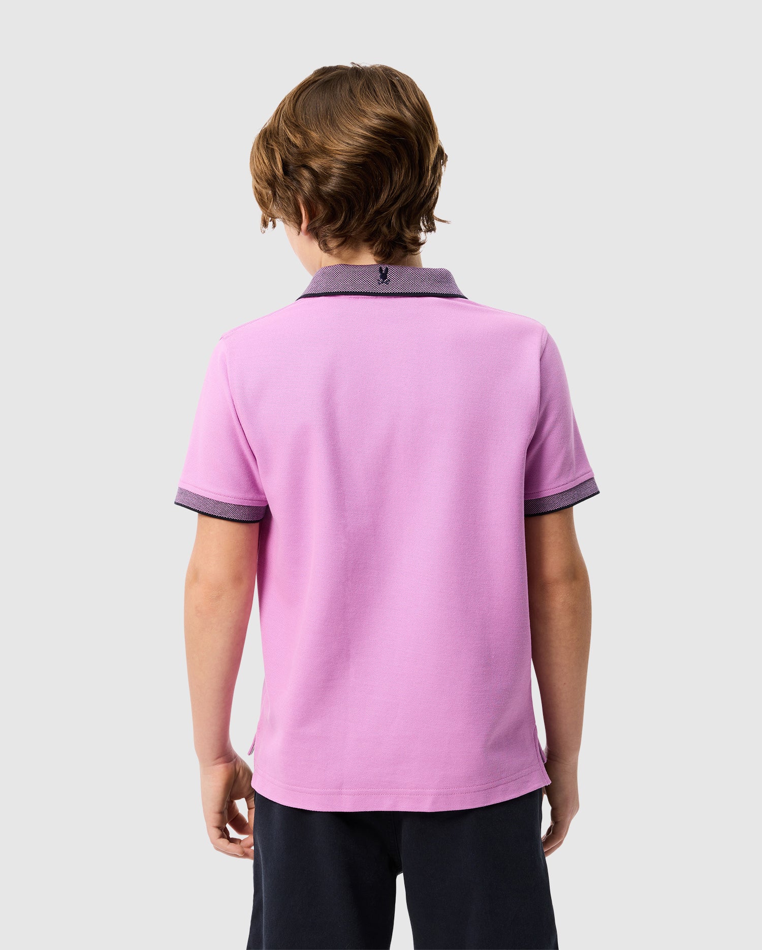 A young person with wavy, light brown hair is shown from behind, wearing a light purple KIDS SOUTHPORT PIQUE POLO SHIRT - B0K263B200 by Psycho Bunny with dark trimming on the collar and sleeves. They are also wearing dark shorts. The background is plain and light grey.