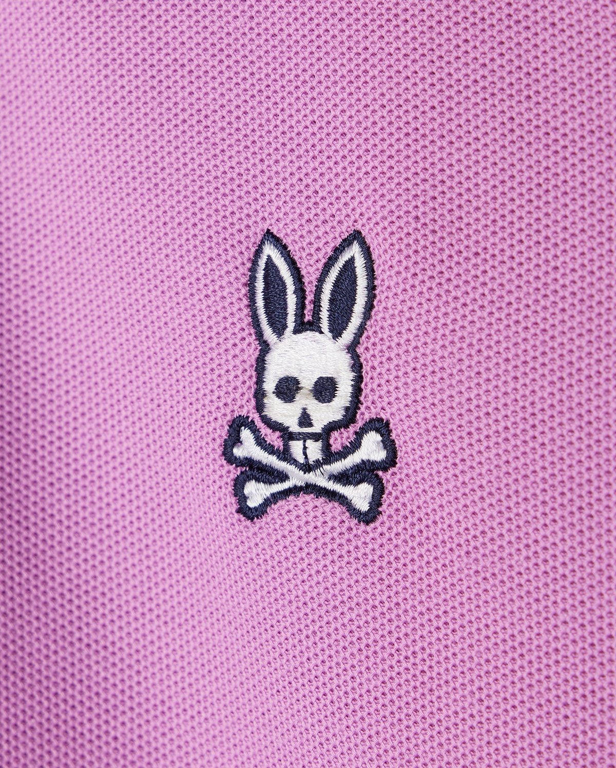 A close-up of pink Psycho Bunny KIDS SOUTHPORT PIQUE POLO SHIRT - B0K263B200 fabric with a small embroidered white bunny skull and crossbones design. The bunny has long ears, black eyes, and black detail on the ears and skull. The fabric, reminiscent of a kids polo, has a textured, mesh-like appearance.