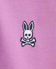 A close-up of pink Psycho Bunny KIDS SOUTHPORT PIQUE POLO SHIRT - B0K263B200 fabric with a small embroidered white bunny skull and crossbones design. The bunny has long ears, black eyes, and black detail on the ears and skull. The fabric, reminiscent of a kids polo, has a textured, mesh-like appearance.